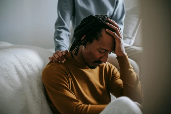 pexels-photo-5700186-5700186 Worried depressed African American male touching head nervously and sitting with eyes closed near supporting wife in bedroom
