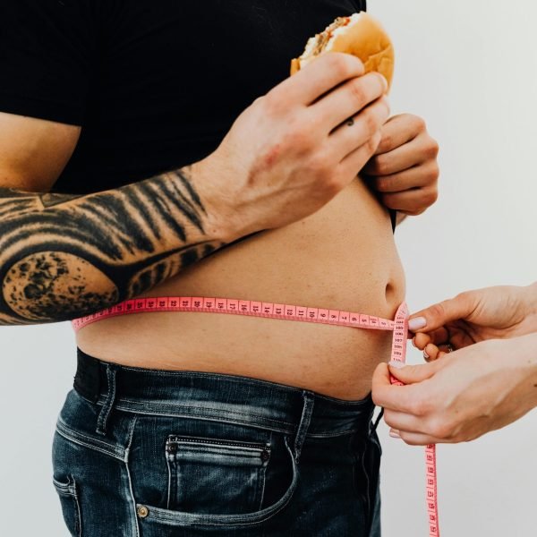 pexels-photo-5714314-5714314 Close-up of a man holding a burger while measuring his waistline with a tape, highlighting unhealthy eating habits.