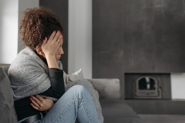 pexels-photo-6753175-6753175 A woman sits on a couch indoors, wrapped in a blanket, holding her head in discomfort.